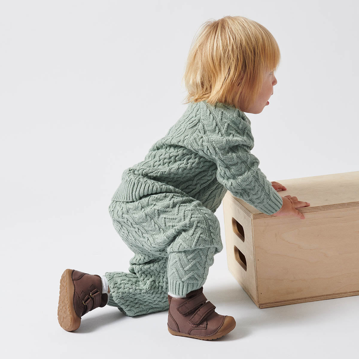 Baby iført strikket sæt i grøn og Bundgaard begyndersko i brun, kravler op ad en træboks – perfekte sko til barnets første skridt.